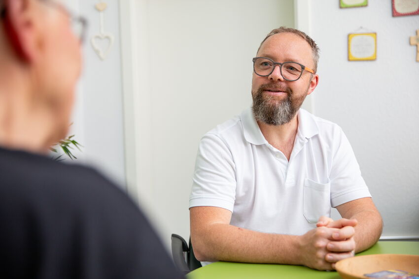 Dr. med. Markus Gläser sitzt lächelnd an einem Tisch und blickt einem Gesprächspartner entgegen. Die Wände sind mit kleinen bunten Bildern dekoriert. Die Stimmung ist offen und zugewandt.