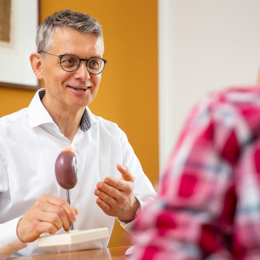 Dr. med. Jörg Schwöbel sitzt an einem Schreibtisch und führt ein Gespräch mit einer Patientin. Er lächelt ihr zu. In der Hand hält er ein Nachbildung einer Niere. Die Szene spielt in einem modern eingerichteten Besprechungsraum.