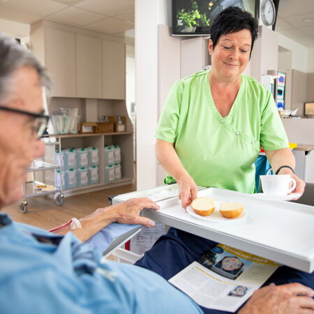 Ein Patient in blauem Dialysekittel sitzt in einem Klinikraum und liest eine Zeitschrift. Eine Pflegekraft in grüner Kleidung reicht ihm ein Tablett mit Getränk und Obst. Die Szene zeigt fürsorgliche Betreuung während einer Dialysebehandlung in angenehmer