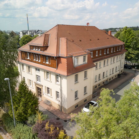 Luftaufnahme des Dialysezentrums Chemnitz mit rotem Ziegeldach und hellen Fassaden. Das Gebäude liegt ruhig inmitten von Bäumen; im Vordergrund steht ein weißes Fahrzeug. Der Himmel ist leicht bewölkt, weitere Gebäude sind im Hintergrund zu erkennen.