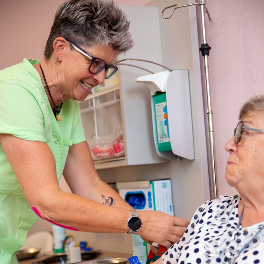 Eine Pflegekraft mit Brille richtet das Handgelenk einer älteren Patientin im Behandlungsstuhl. Die Patientin ist mit einer gepunkteten Bluse bekleidet. Die Szene zeigt eine ruhige Behandlungssituation in einem klinischen Raum.