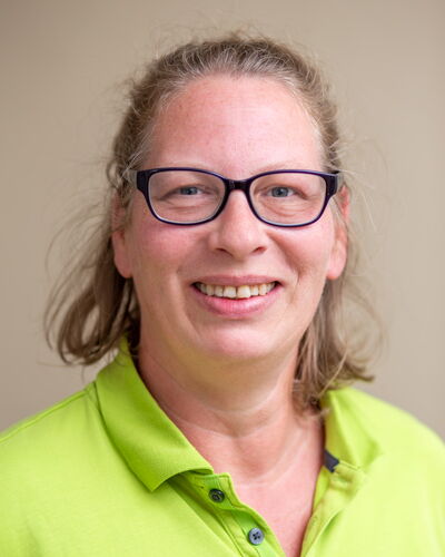 Praxismanagerin Juliane Scheunert steht vor einer hellen Wand und lächelt. Die offene Haltung unterstützt ihren verantwortungsvollen Einsatz im administrativen Bereich des Dialysezentrums.