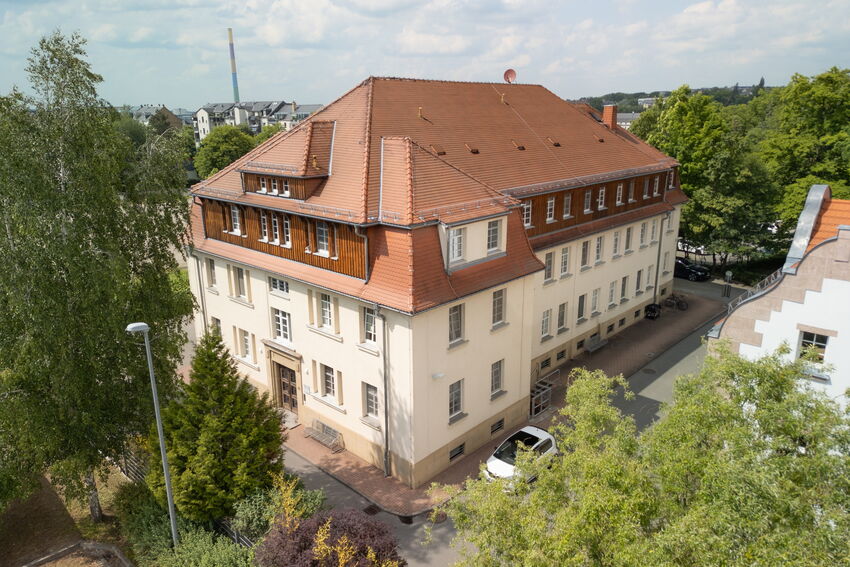 Luftaufnahme des Dialysezentrums Chemnitz mit rotem Ziegeldach und hellen Fassaden. Das Gebäude liegt ruhig inmitten von Bäumen; im Vordergrund steht ein weißes Fahrzeug. Der Himmel ist leicht bewölkt, weitere Gebäude sind im Hintergrund zu erkennen.